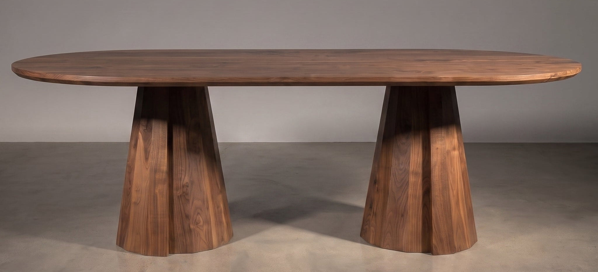 An oval wooden dining table with a rich medium-brown finish, supported by two sculptural, flared pedestal bases, set against a neutral gray studio background.