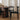 A dark wood dining table with a cylindrical base and black chairs, set in a bright room with sheer white curtains and a light wood herringbone floor.